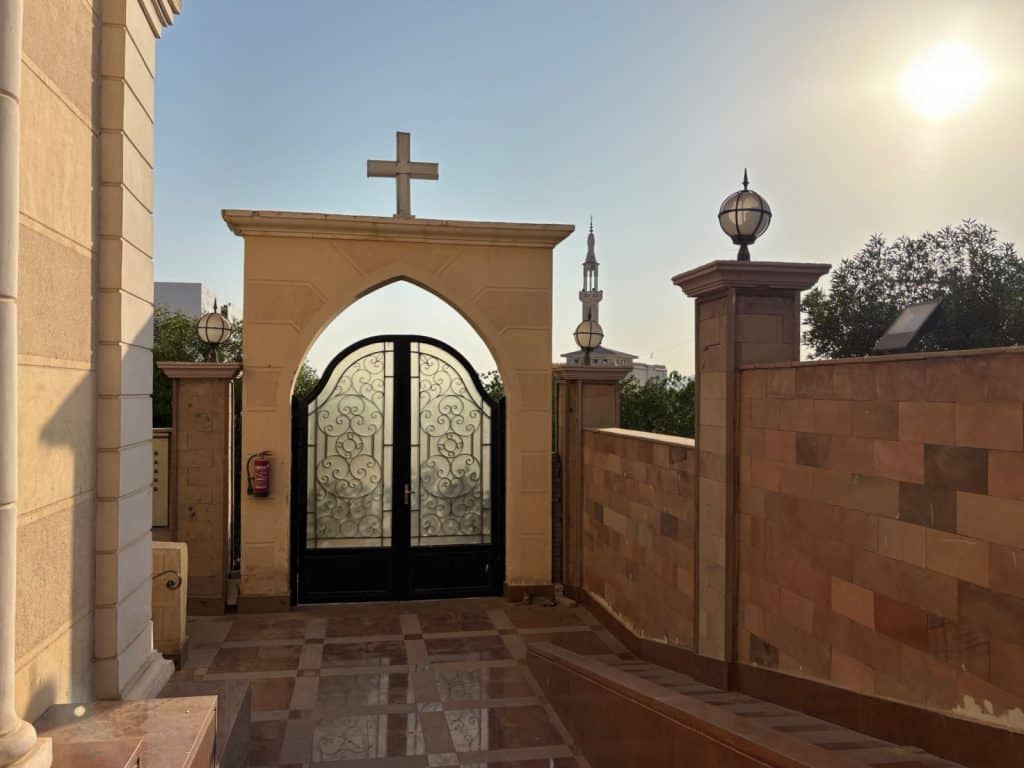 Ein gemauertes Tor mit einem christlichen Kreuz an der Spitze führt auf das Gelände einer Kirche in Sharm El Sheikh, während im Hintergrund das Minarett einer Moschee unter der hellen Sonne sichtbar ist.