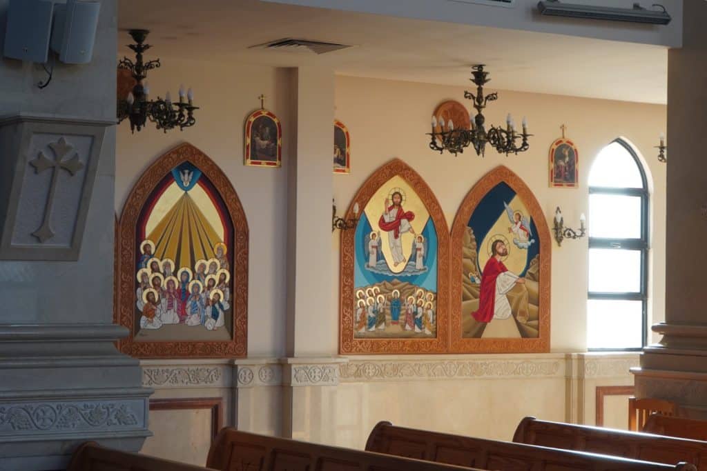 Innenansicht einer Kirche in Sharm El Sheikh mit kunstvollen Wandbildern in Spitzbogenrahmen, die biblische Szenen im byzantinischen Stil darstellen, sowie hölzernen Kirchenbänken und dekorativen Kronleuchtern.