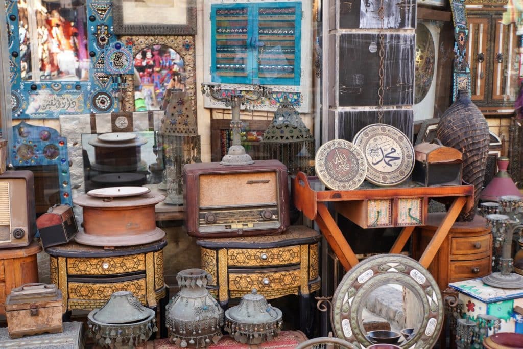 Ein vollgestellter Ladenstand auf dem Souk in Scharm El-Scheich präsentiert eine Vielzahl an Antiquitäten, darunter verzierte Holzkommoden, alte Radios, kunstvolle Metallgefäße und dekorative Teller mit arabischer Kalligrafie. Im Hintergrund ergänzen bunte Spiegelrahmen und traditionelle Laternen das dichte Angebot an ägyptischem Kunsthandwerk.