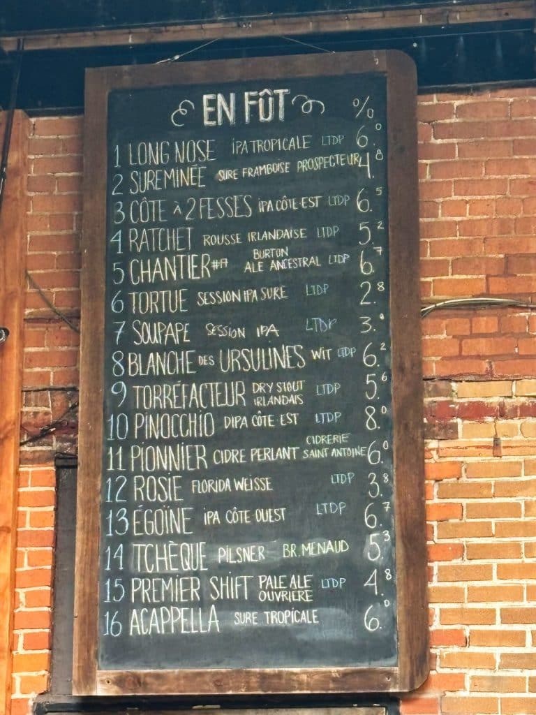 Eine hölzerne Menütafel an einer Backsteinwand listet 16 verschiedene Biersorten der Brauerei Le Temps d'une Pinte in Trois-Rivières auf, inklusive Namen, Stilen und Alkoholgehalt. Jede Sorte ist nummeriert und in weißer Kreide handschriftlich unter der Überschrift „EN FÛT“ (vom Fass) aufgeführt.