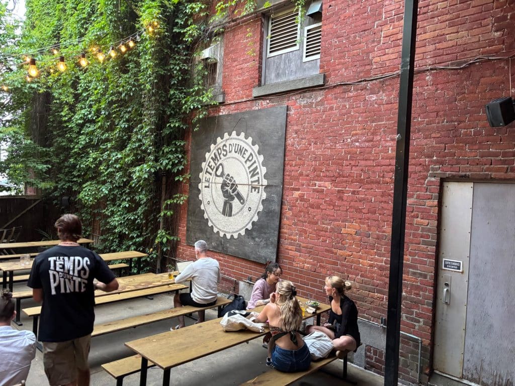 Gäste sitzen an Holztischen im gemütlichen Außenbereich der Brauerei Le Temps d'une Pinte in Trois-Rivières, der von einer backsteinernen Wand mit großem Logo und einer üppig bewachsenen Efeuwand eingerahmt wird.