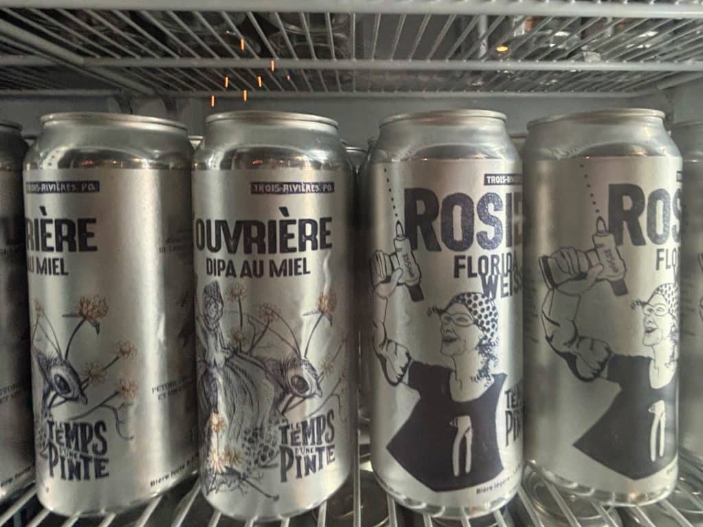 Vier silberne Bierdosen der Brauerei Le Temps d'une Pinte aus Trois-Rivières stehen nebeneinander in einem Kühlschrankregal, bedruckt mit schwarzen Illustrationen und den Namen „Ouvrière“ und „Rosie“.