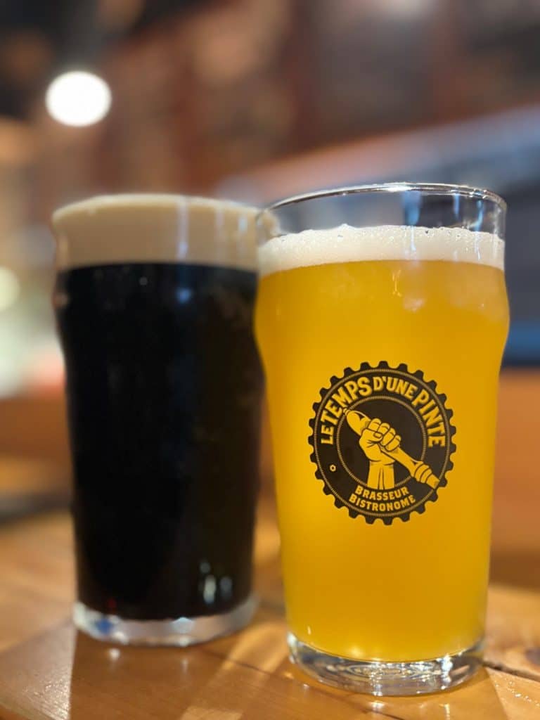 Zwei Gläser Bier der Brauerei Le Temps d'une Pinte in Trois-Rivières stehen auf einem Holztisch, wobei ein helles, trübes Bier im Vordergrund und ein dunkles Bier mit cremiger Schaumkrone im Hintergrund zu sehen ist.