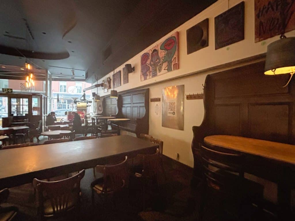 Innenansicht der Trois-Rivières-Brauerei mit rustikalen Holztischen, dunklen Wandpaneelen und farbenfrohen Kunstwerken an der Wand unter einer hohen, dunklen Decke.