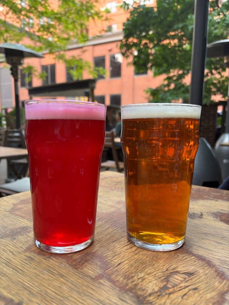 Zwei Gläser Bier der Trois Rivières Brauerei, ein leuchtend rotes Sauerbier und ein goldfarbenes Lagerbier, stehen auf einem Holztisch im Außenbereich.