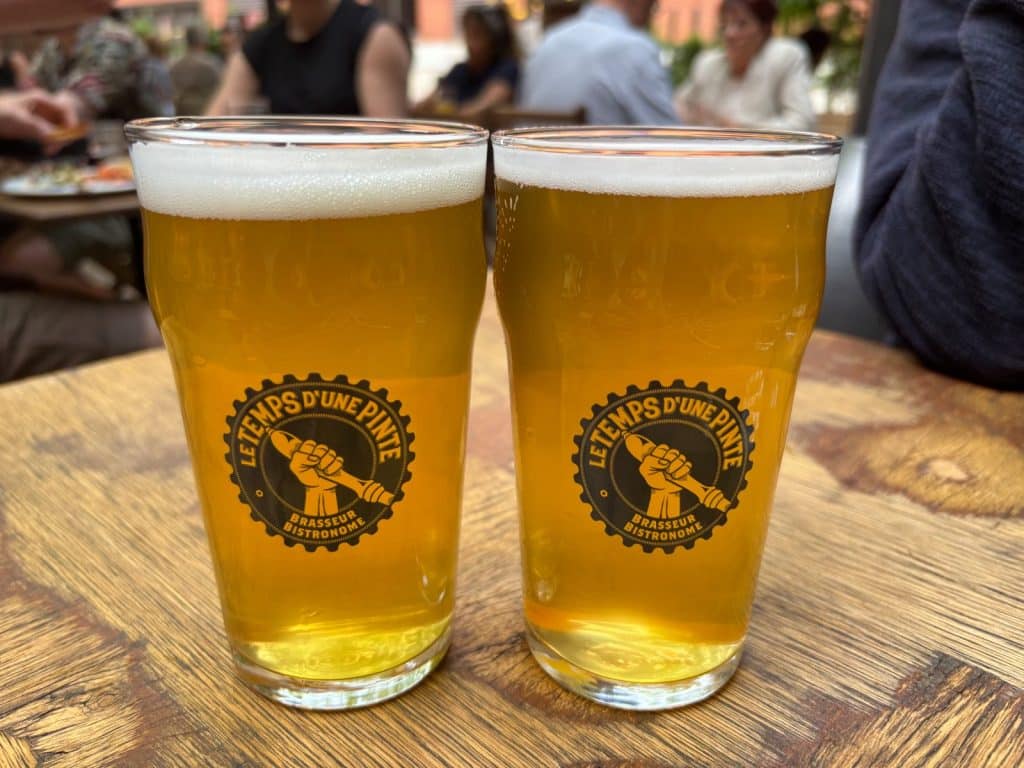 Zwei Gläser helles Bier mit weißer Schaumkrone stehen auf einem Holztisch in der Brauerei Le Temps d'une Pinte in Trois-Rivières.