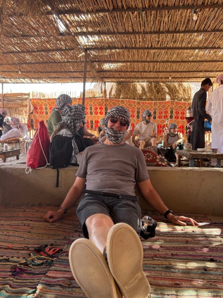 Ein Mann mit Sonnenbrille und schwarz-weiß kariertem Kopftuch lehnt entspannt in einem traditionellen ägyptischen Zelt auf einem bunten Teppich.