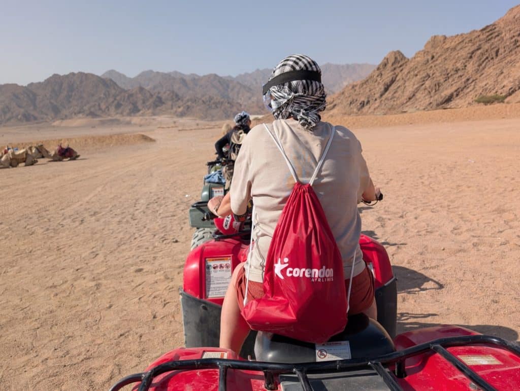 Eine Gruppe von Menschen fährt auf roten Quads durch die ägyptische Wüste, wobei im Vordergrund eine Person mit einem rot-weißen Rucksack und einem schwarz-weiß karierten Kopftuch zu sehen ist.