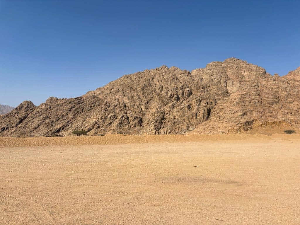 Ausflugtipps Sharm El Sheikh: Eine weite, sandige Wüstenlandschaft in Ägypten unter einem klaren blauen Himmel, im Hintergrund begrenzt von einem massiven, zerklüfteten Felsgebirge.