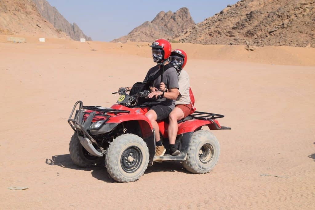 Zwei Personen mit roten Helmen und Schutzmasken fahren gemeinsam auf einem roten Quad durch eine weitläufige ägyptische Wüstenlandschaft vor einer felsigen Bergkulisse.