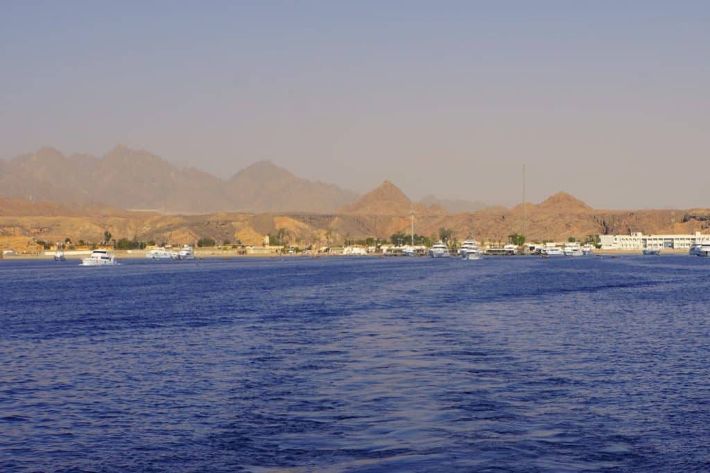 Blick vom tiefblauen Meer auf die ägyptische Küste mit zahlreichen weißen Schiffen vor einer kargen, bergigen Wüstenlandschaft unter dunstigem Himmel.