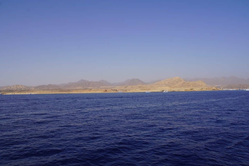 Blick von einem Schiff auf die weit entfernte, karge Wüstenküste Ägyptens mit schroffen Bergen unter einem klaren blauen Himmel.