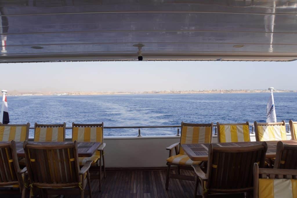Ausflugtipps Sharm El Sheikh: Blick vom überdachten Deck eines Schiffes in Ägypten auf das blaue Meer und die ferne Küste, mit hölzernen Tischen und gelb-weiß gestreiften Sitzplätzen im Vordergrund.