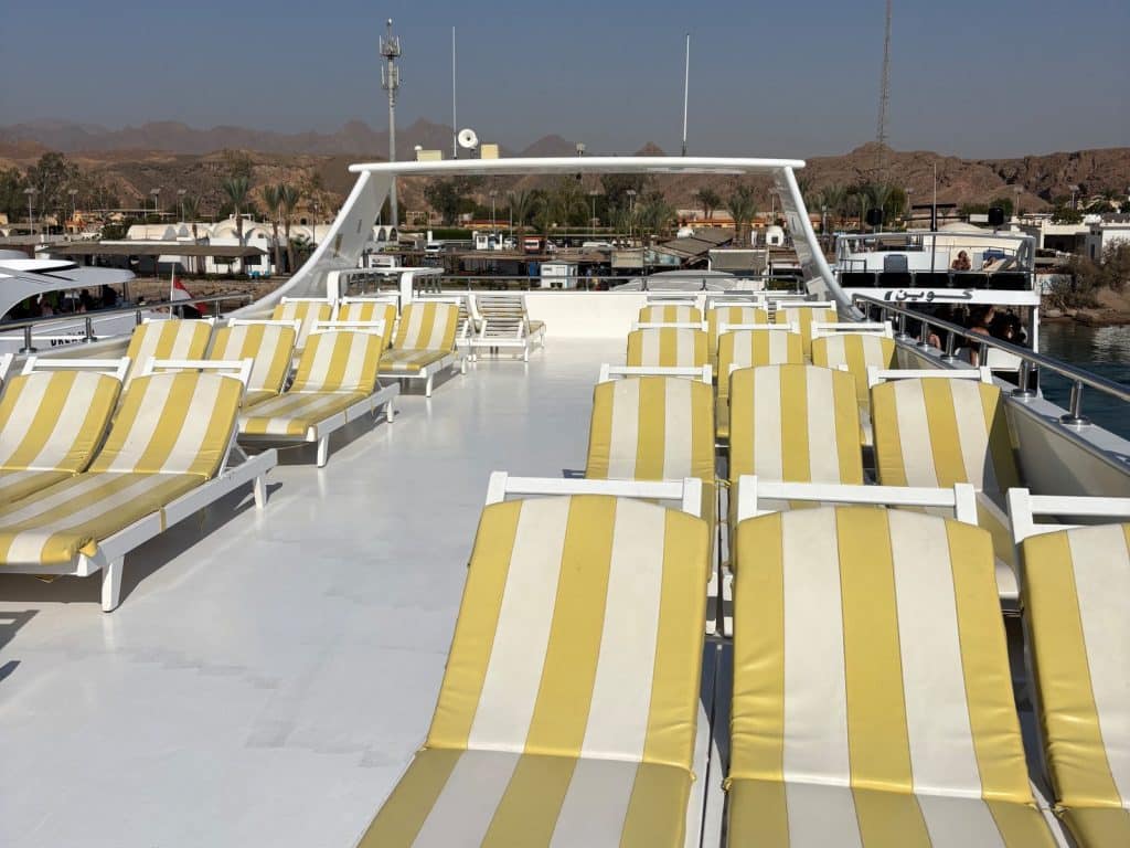 Ausflugtipps Sharm El Sheikh: Das obere Sonnendeck eines Schiffes in Ägypten mit zahlreichen weiß-gelb gestreiften Liegestühlen vor einer Kulisse aus Bergen und Küstengebäuden.