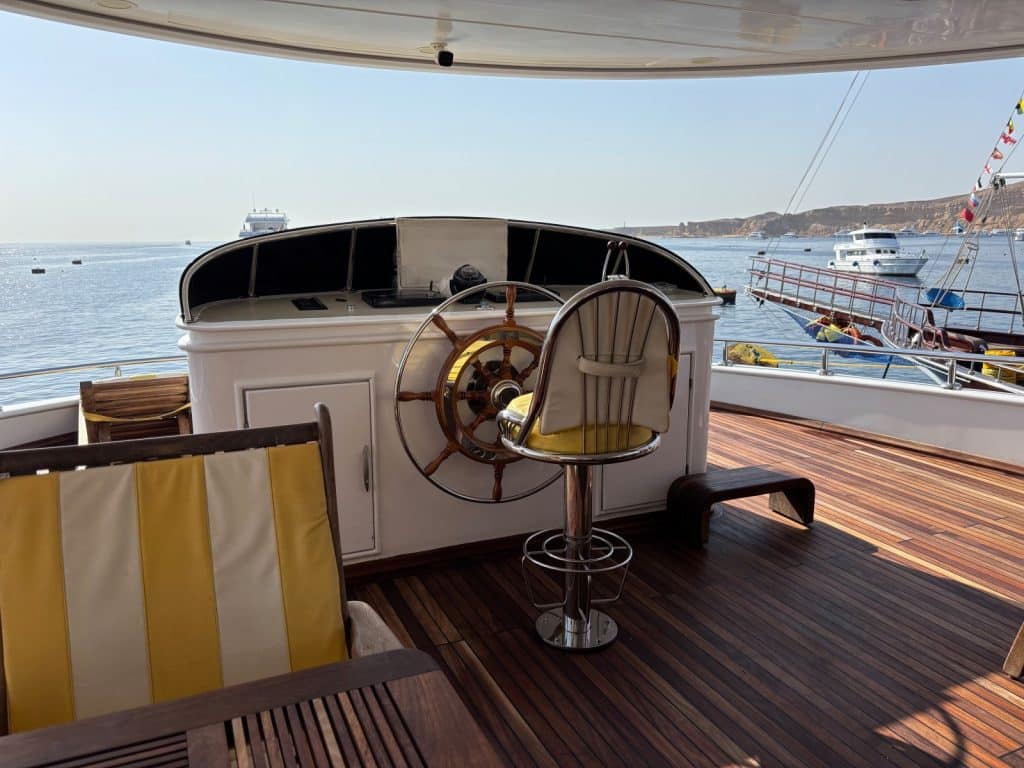 Blick vom hölzernen Deck eines Schiffes in Ägypten auf ein klassisches hölzernes Steuerrad und einen gelb-weißen Kapitänssessel vor einer Meereskulisse mit weiteren Booten.