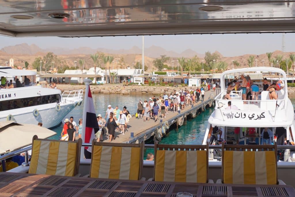 Blick von einem Schiff auf eine Anlegestelle in Ägypten, auf der zahlreiche Touristen über einen Steg zu bereitstehenden Ausflugsbooten laufen. Im Vordergrund sind gestreifte Sitzpolster und ein Holztisch an Deck zu sehen, während im Hintergrund Palmen und karge Berge unter klarem Himmel aufragen.