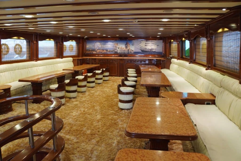 Das Bild zeigt das luxuriös ausgestattete Unterdeck eines ägyptischen Schiffes mit dunklen Holzpaneelen, einer hellen, gepolsterten Sitzbank entlang der Wände und mehreren polsterten Hockern an Marmortischen auf einem flauschigen, beigefarbenen Teppich. Den hinteren Abschluss bildet eine beleuchtete Wand mit maritimen Panoramabildern unter einer niedrigen Decke mit eingelassenen Spots.