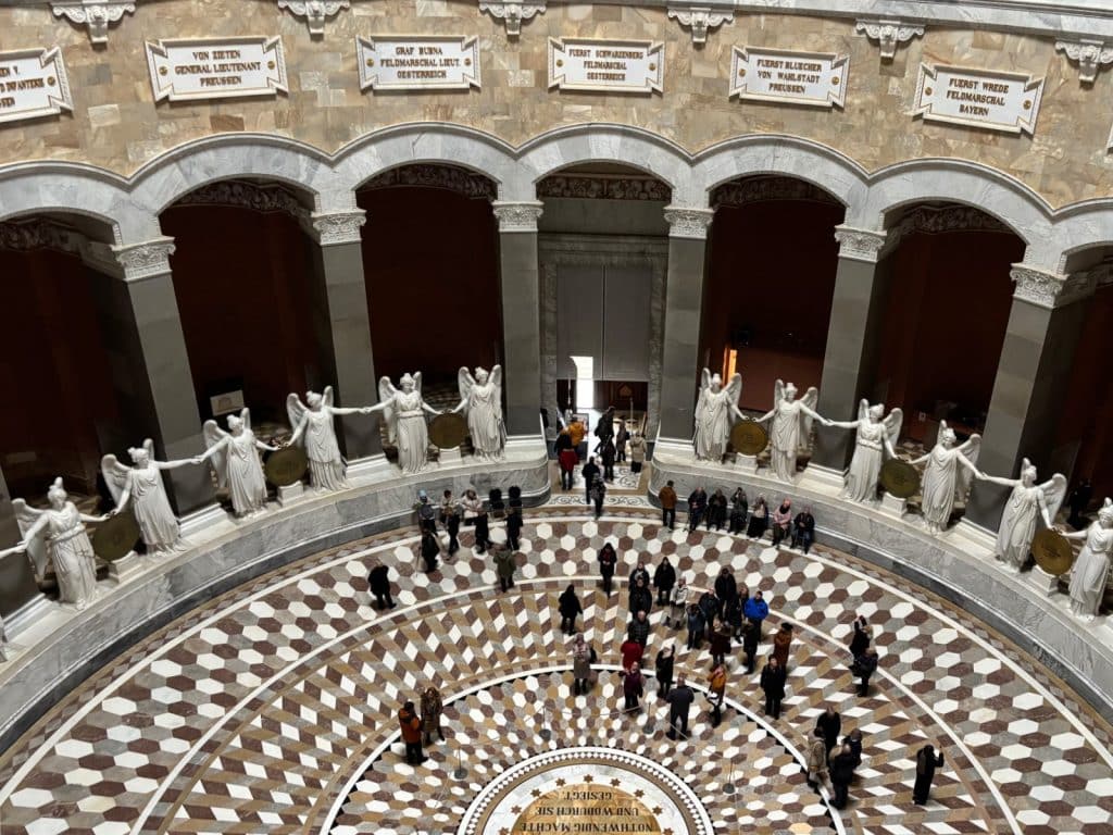 Blick von der Galerie in den kreisförmigen Innenraum der Befreiungshalle, in dem zahlreiche weiße Marmorstatuen von Siegesgöttinnen Hand in Hand vor hohen Arkaden stehen und Besucher über den geometrisch gemusterten Mosaikboden wandeln.