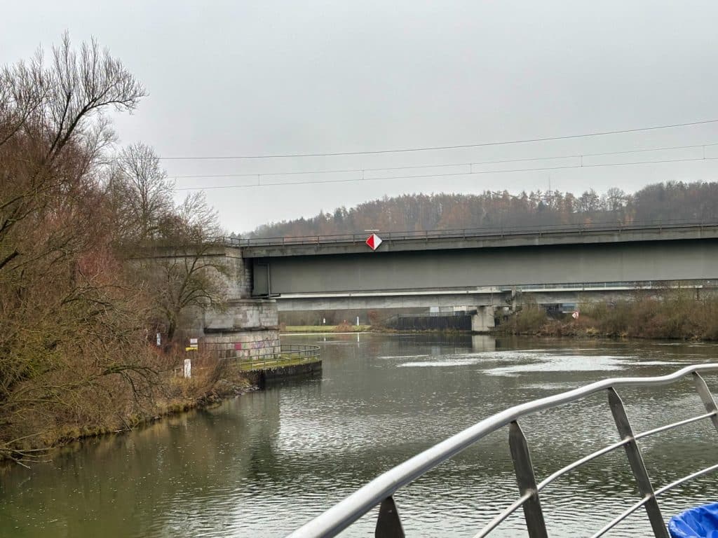 Blick von einem Schiff auf die niedrige Brücke, die über einen ruhigen Fluss führt und von herbstlichen Bäumen gesäumt wird.