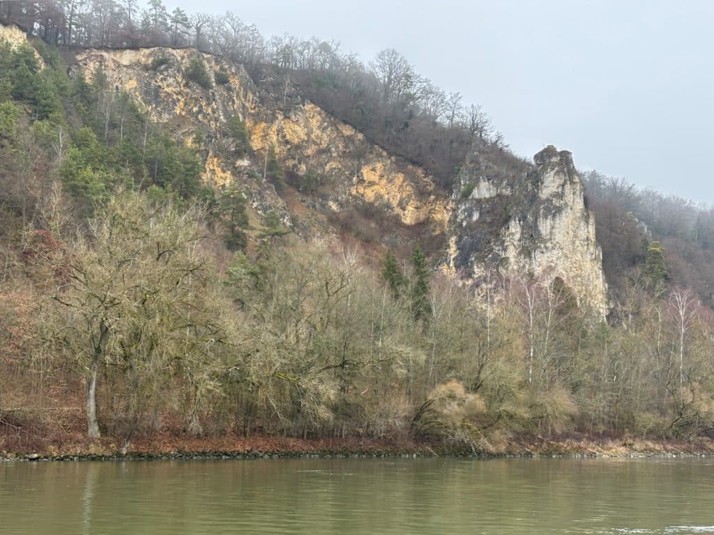 Die Uferfelsen ragen als steile, helle Kalksteinwände hinter einer dichten Reihe herbstlicher Bäume am Ufer eines ruhig dahinfließenden Flusses empor.