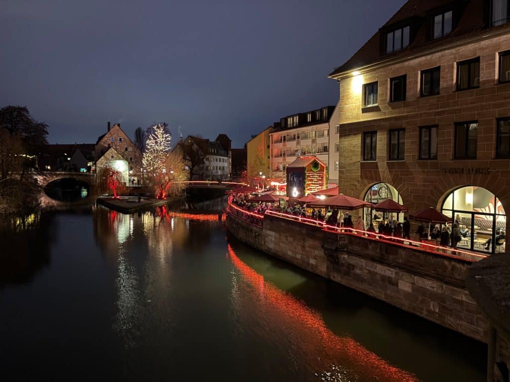 Abendliche Ansicht der weihnachtlich beleuchteten Nürnberger Altstadt mit einer Uferpromenade an der Pegnitz, an der Menschen unter roten Schirmen vor einem Starbucks-Café verweilen. Ein hell erleuchteter Weihnachtsbaum und festliche Lichterketten spiegeln sich stimmungsvoll im dunklen Wasser des Flusses.