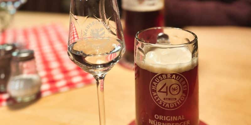 Ein Glas Schnaps steht neben einem Krug mit Original Nürnberger Rotbier der Hausbrauerei Altstadthof auf einem Holztisch.