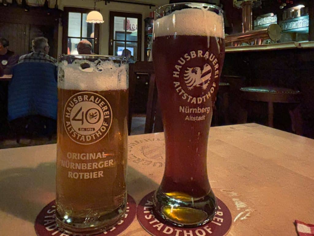Zwei Gläser Nürnberger Rotbier der Hausbrauerei Altstadthof stehen auf einem Holztisch in einer gemütlichen Brauhaus-Atmosphäre. Das linke Glas ist ein Krug mit dem Aufdruck „40 Jahre“, während das rechte ein hohes, schlankes Weizenglas mit dem Brauerei-Logo ist.