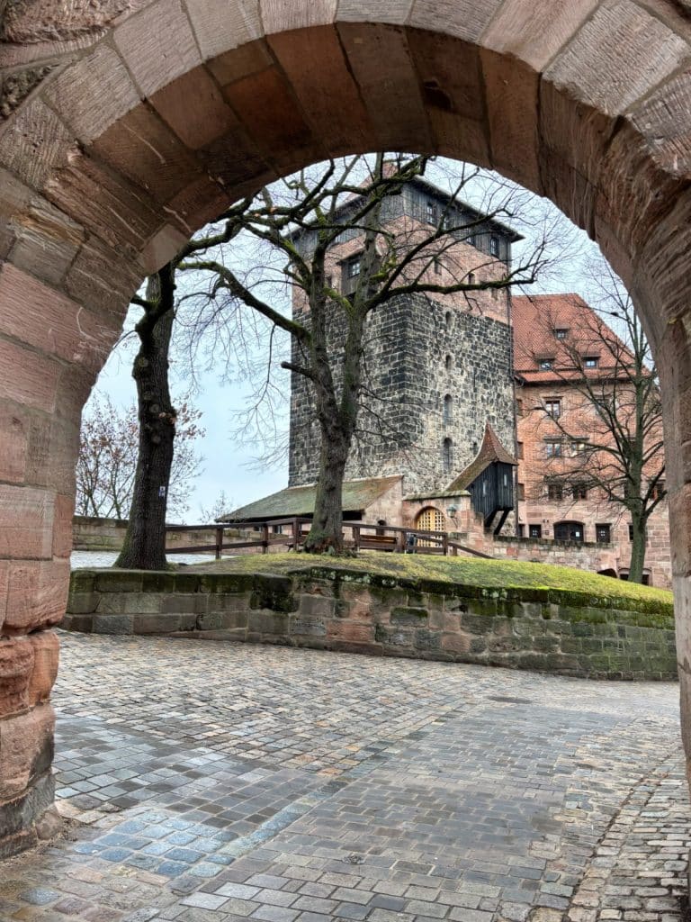 Blick durch einen steinernen Torbogen auf den mittelalterlichen Burgturm der Nürnberger Burg, umgeben von kahlen Bäumen und gepflasterten Wegen.