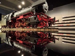 Eine historische Dampflokomotive mit roten Rädern steht auf Schienen im DB Museum Nürnberg und spiegelt sich perfekt in der glänzenden, schwarzen Bodenfläche. Im Hintergrund sind beleuchtete Vitrinen mit zahlreichen Miniatur-Modellzügen an der Wand zu sehen.