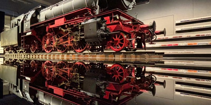 Eine historische Dampflokomotive mit roten Rädern steht auf Schienen im DB Museum Nürnberg und spiegelt sich perfekt in der glänzenden, schwarzen Bodenfläche. Im Hintergrund sind beleuchtete Vitrinen mit zahlreichen Miniatur-Modellzügen an der Wand zu sehen.
