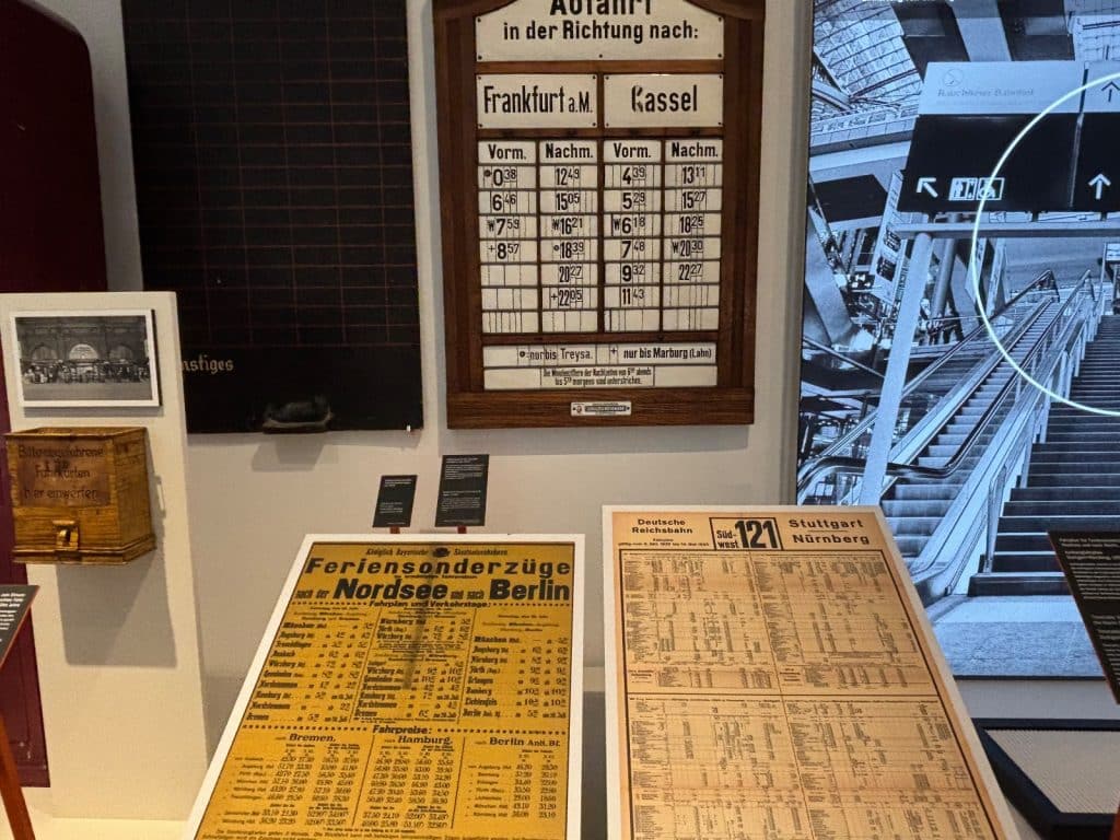 Historische Abfahrtstafeln und Fahrpläne der Deutschen Reichsbahn in einer Ausstellung des DB Museums in Nürnberg, darunter eine hölzerne Wandtafel mit Zugverbindungen nach Frankfurt am Main und Kassel.