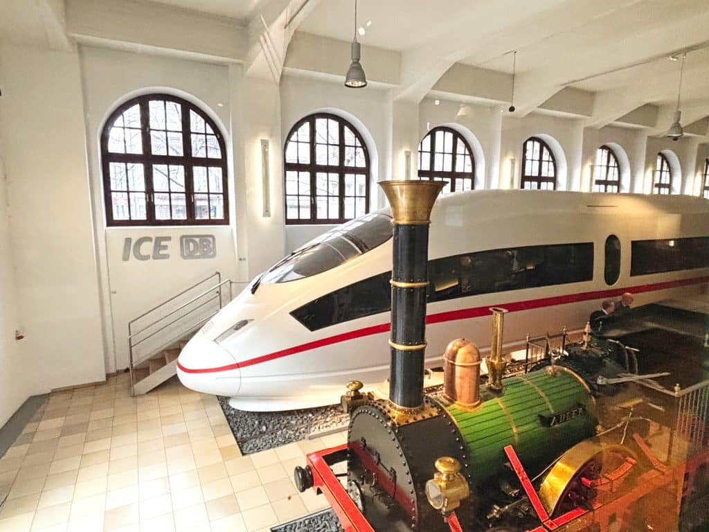 Im DB Museum Nürnberg steht ein moderner, weißer ICE-Hochgeschwindigkeitszug direkt hinter dem historischen Nachbau der ersten deutschen Dampflokomotive „Adler“.