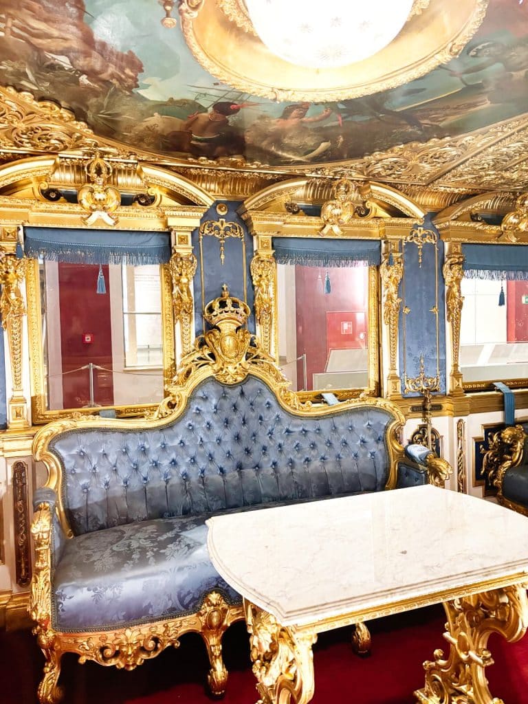 Ein prunkvolles Abteil im Königszug von Ludwig II. im DB Museum Nürnberg mit einem blau gepolsterten Sofa, einem Marmortisch und reichhaltigen Goldverzierungen an Wänden und Decke. Die Decke ist zudem mit einem kunstvollen Gemälde und einer zentralen Leuchte gestaltet.