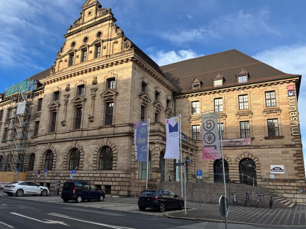 Außenansicht des historischen Gebäudes des DB Museums in Nürnberg mit seiner markanten Sandsteinfassade, davor geparkten Autos und Fahnenmasten unter blauem Himmel.