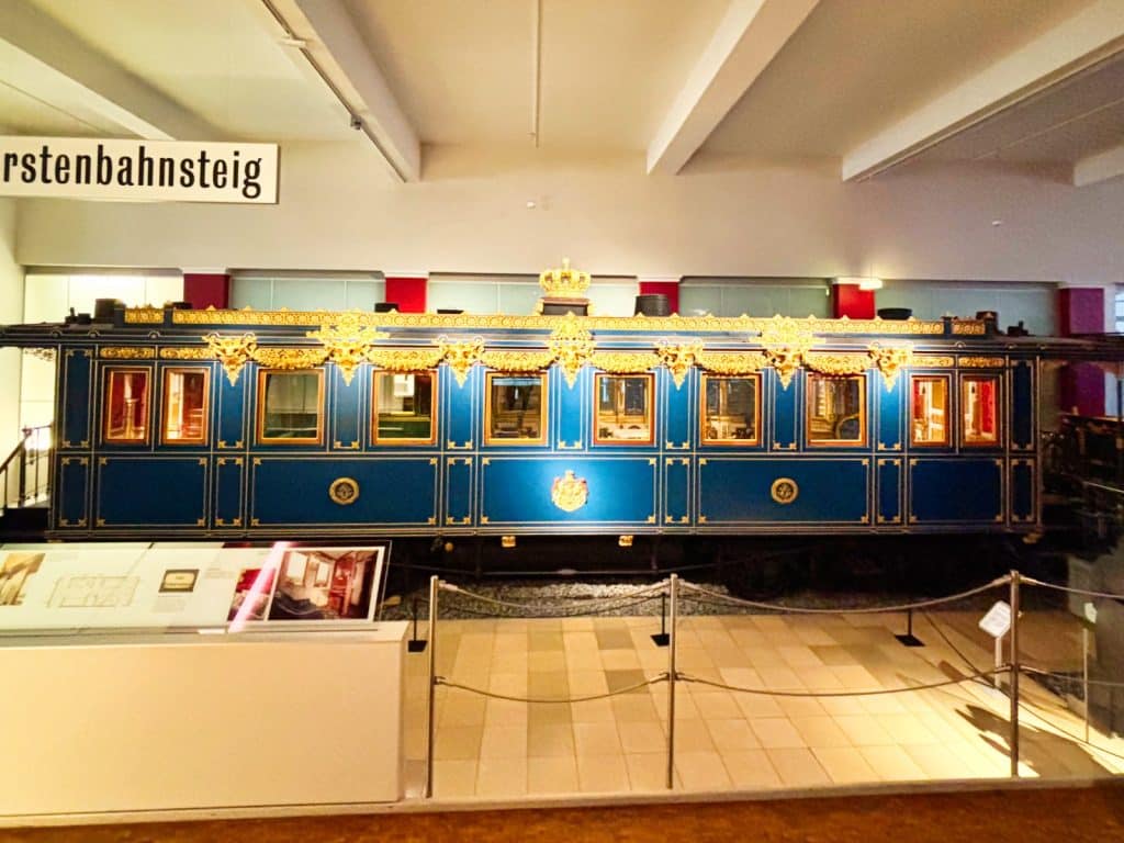 Die Außenansicht eines prunkvollen, blau lackierten Eisenbahnwaggons des Königszugs von König Ludwig II., verziert mit goldenen Ornamenten, Wappen und einer Krone auf dem Dach.