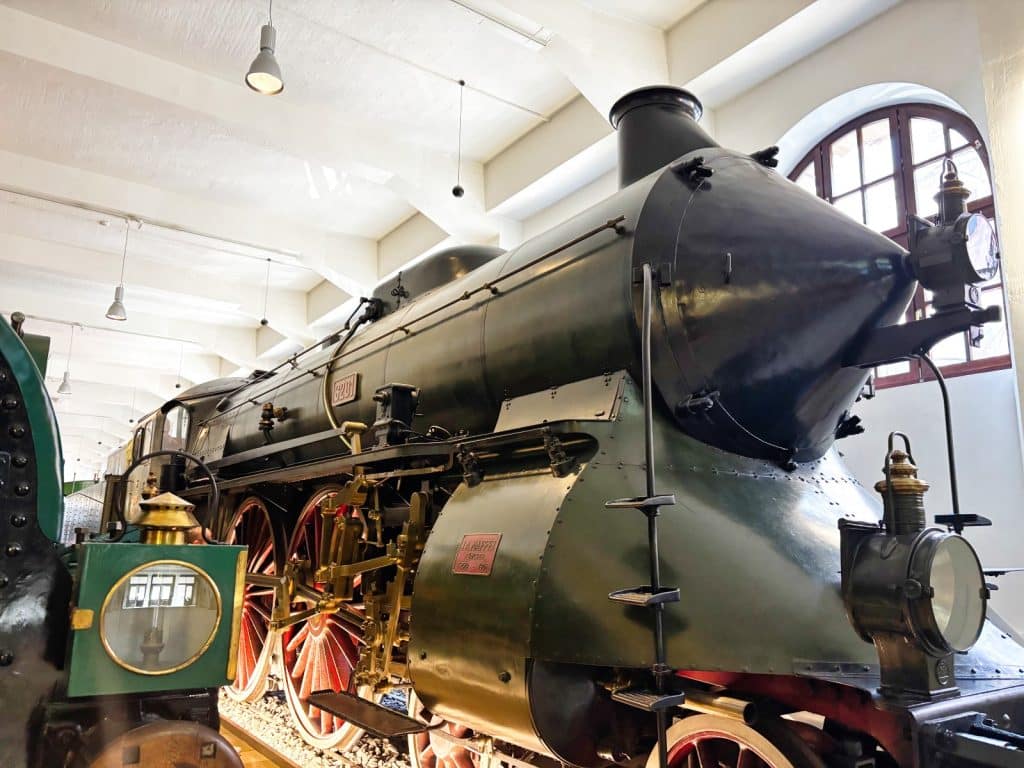 Eine historische Dampflokomotive mit markanter kegelförmiger Front und großen roten Speichenrädern steht in einer Ausstellungshalle des DB Museums in Nürnberg.