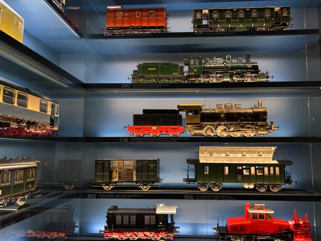 Eine Glasvitrine präsentiert verschiedene historische Modelleisenbahnen auf mehreren Ebenen, darunter detailgetreue Dampflokomotiven, Personenwagen und Güterwaggons in unterschiedlichen Farben und Bauformen.