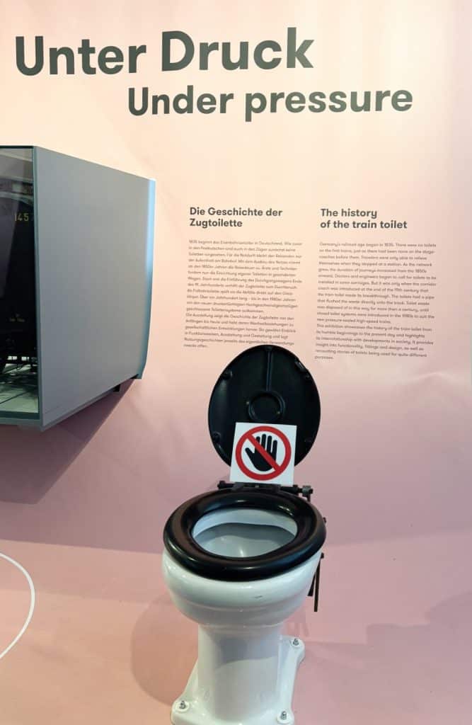 Ein historisches Toilettenbecken aus der Ausstellung „Unter Druck – Die Geschichte der Zugtoilette“ im DB Museum Nürnberg steht vor einer Informationstafel an einer rosa Wand. Das Exponat hat einen schwarzen Klodeckel mit einem „Nicht berühren“-Schild und zeigt die Entwicklung der sanitären Anlagen in Zügen.