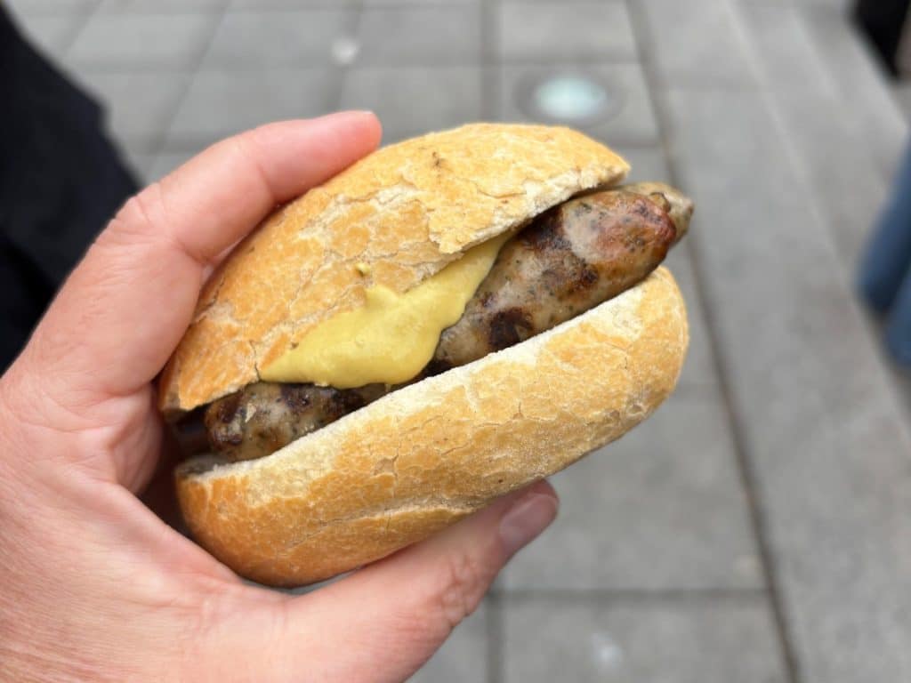 Eine Hand hält ein „Drei im Weckla“ – drei kleine Nürnberger Rostbratwürste mit Senf in einem knusprigen Brötchen vor einem gepflasterten Hintergrund.