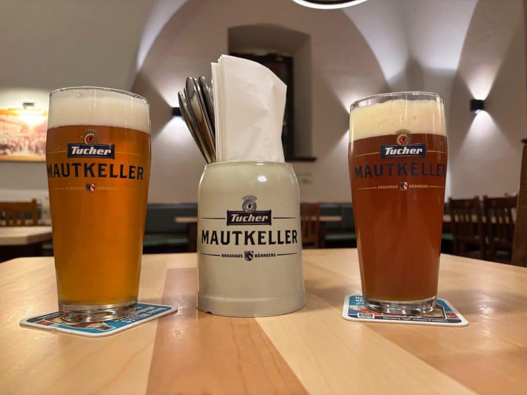 Zwei Gläser Tucher-Bier und ein Krug mit Besteck stehen auf einem Holztisch im Mautkeller in Nürnberg.