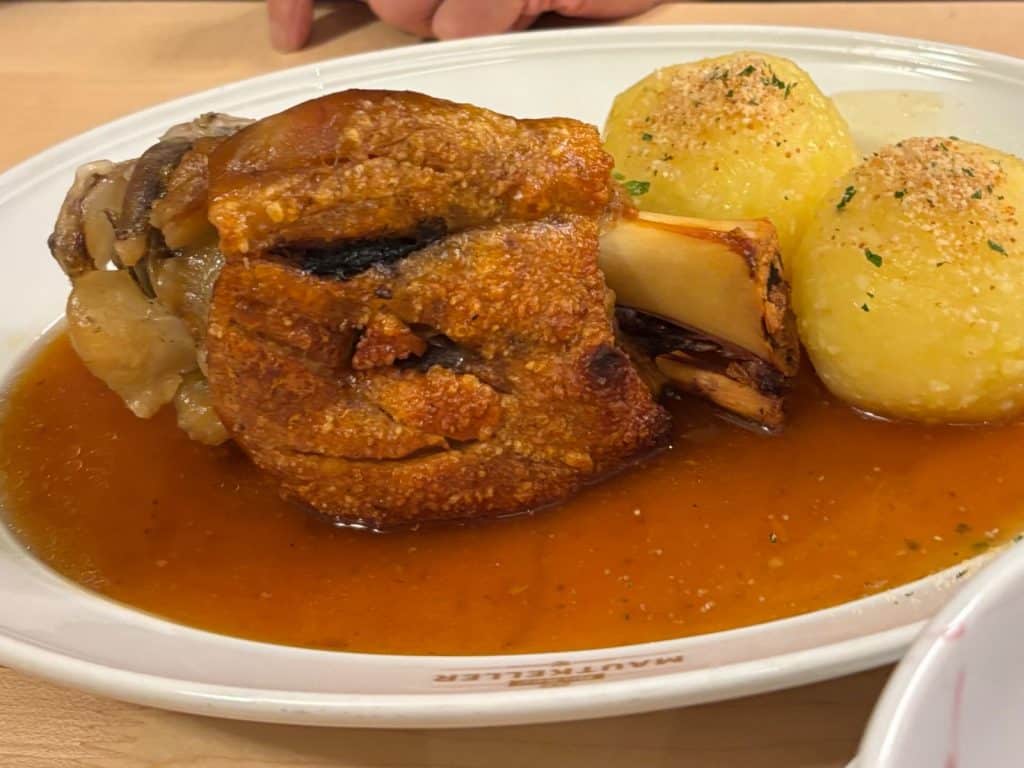Eine knusprig gebratene Schweinshaxe im Mautkeller Nürnberg, serviert mit zwei Kartoffelklößen in einer dunklen Bratensauce auf einem weißen Teller.