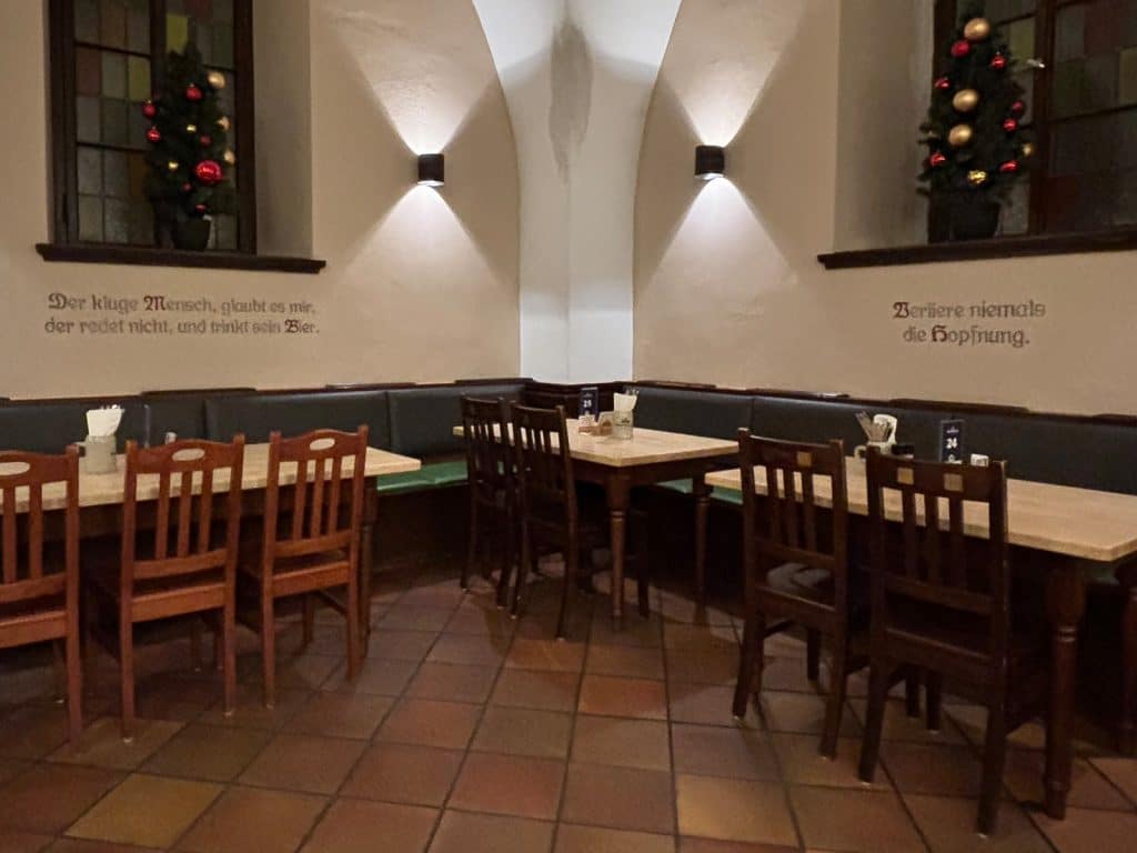 Rustikale Sitzecke im Nürnberger Mautkeller mit Holztischen, Stühlen und Wandsprüchen unter kleinen Weihnachtsbäumen in den Fensternischen.