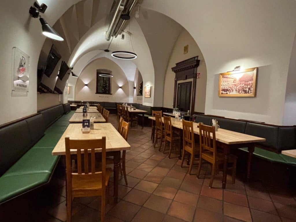 Der Innenraum des Nürnberger Mautkellers zeigt einen langen Gastraum mit gewölbten Decken, rustikalen Holztischen, Stühlen und grünen Sitzbänken auf einem Terrakottaboden. Moderne Ring-Hängeleuchten und Wandstrahler beleuchten die Szenerie, während historische Bilder an den Wänden für ein traditionelles Ambiente sorgen.