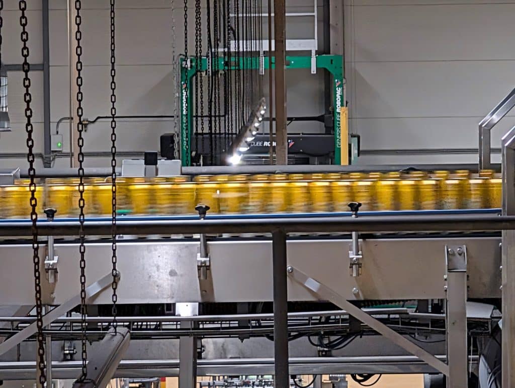 In einer Brauerei in Pilsen bewegen sich goldgelbe Dosen in schneller Folge auf einem industriellen Förderband durch die Abfüllanlage. Im Hintergrund sind komplexe Maschinenstrukturen, Kettenzüge und das grüne Gestell einer Verpackungsmaschine zu sehen.