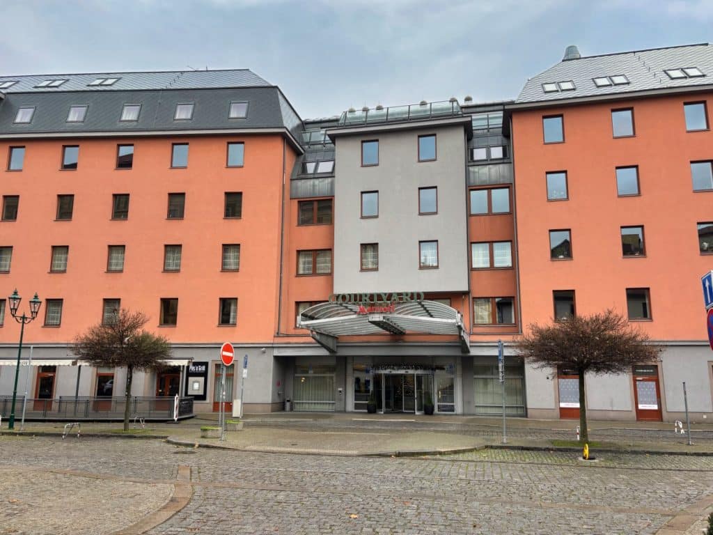 Außenansicht des Courtyard by Marriott Hotels in Pilsen mit seiner markanten lachsfarbenen Fassade, einem modernen grauen Eingangsbereich und einem gepflasterten Vorplatz.