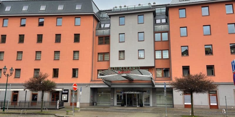 Außenansicht des Courtyard by Marriott Hotels in Pilsen mit seiner markanten lachsfarbenen Fassade, einem modernen grauen Eingangsbereich und einem gepflasterten Vorplatz.