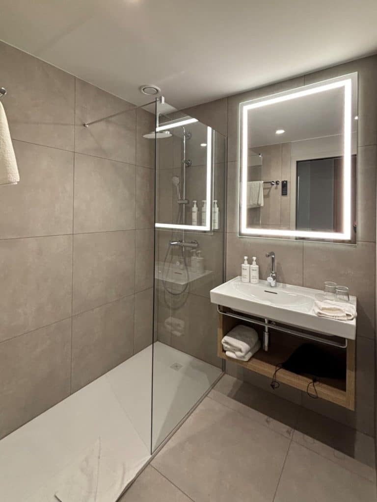 Modernes Badezimmer im Courtyard by Marriott Pilsen mit einer bodengleichen Glasdusche, einem weißen Waschbecken mit beleuchtetem Spiegel und großformatigen, beigefarbenen Wandfliesen.