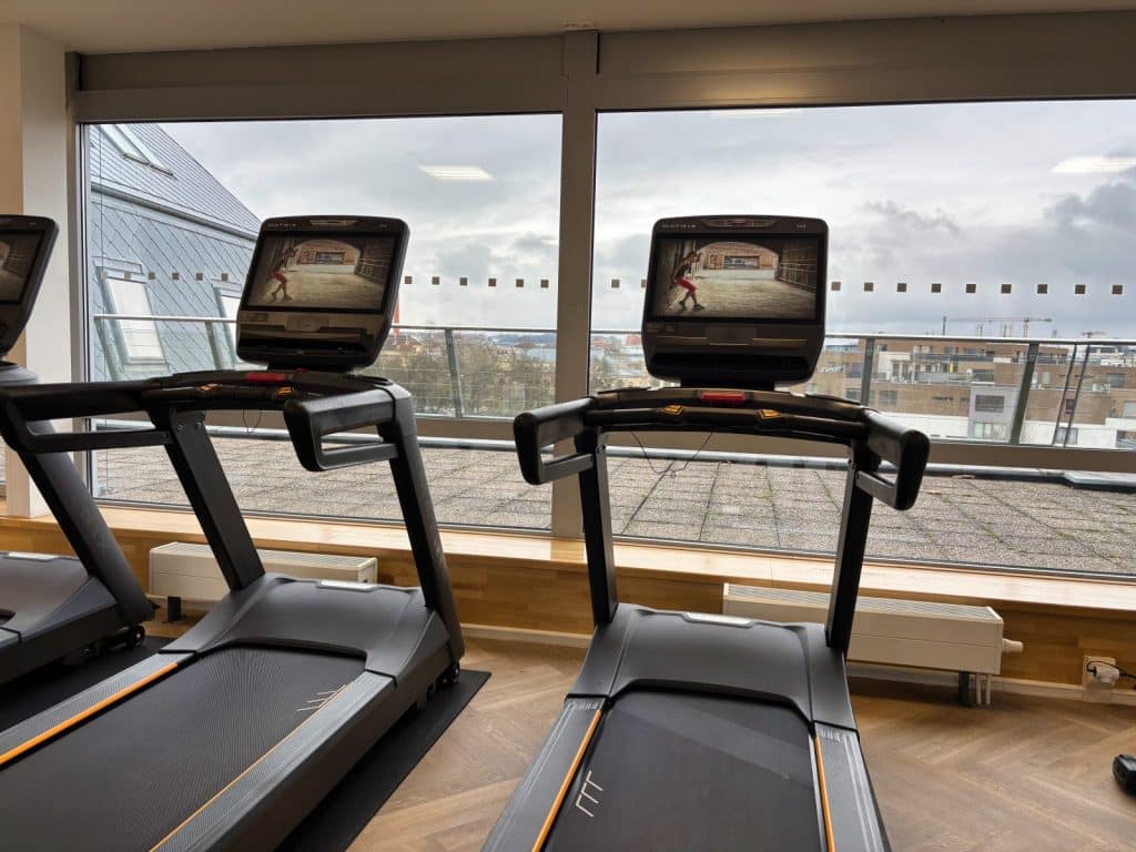 Zwei moderne Laufbänder mit Bildschirmen stehen im Fitnesscenter des Pilsen Marriott Hotels vor einer großen Fensterfront mit Blick auf die Stadt.