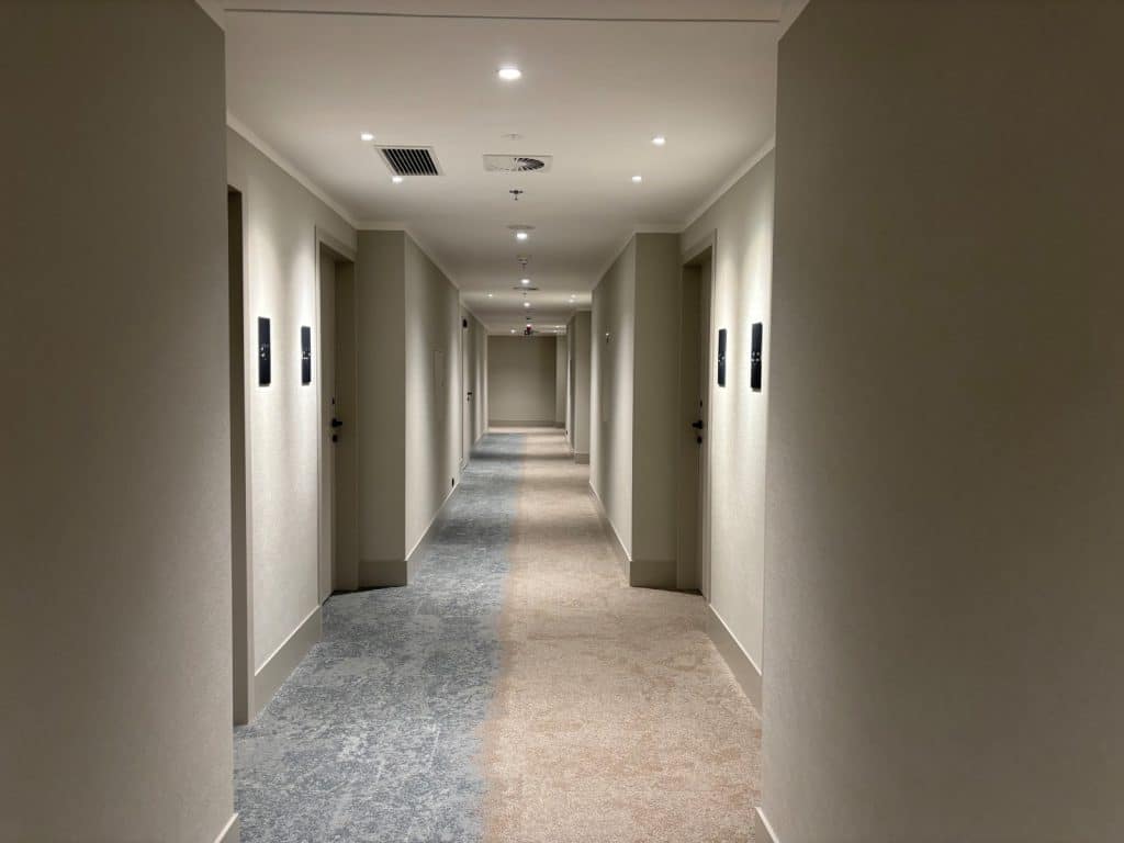 Ein modern gestalteter Hotelgang im Courtyard by Marriott Pilsen mit hellen Wänden, mehreren Zimmertüren und einem zweifarbigen Teppichboden in Blau und Beige, der den Flur optisch in zwei Hälften teilt.