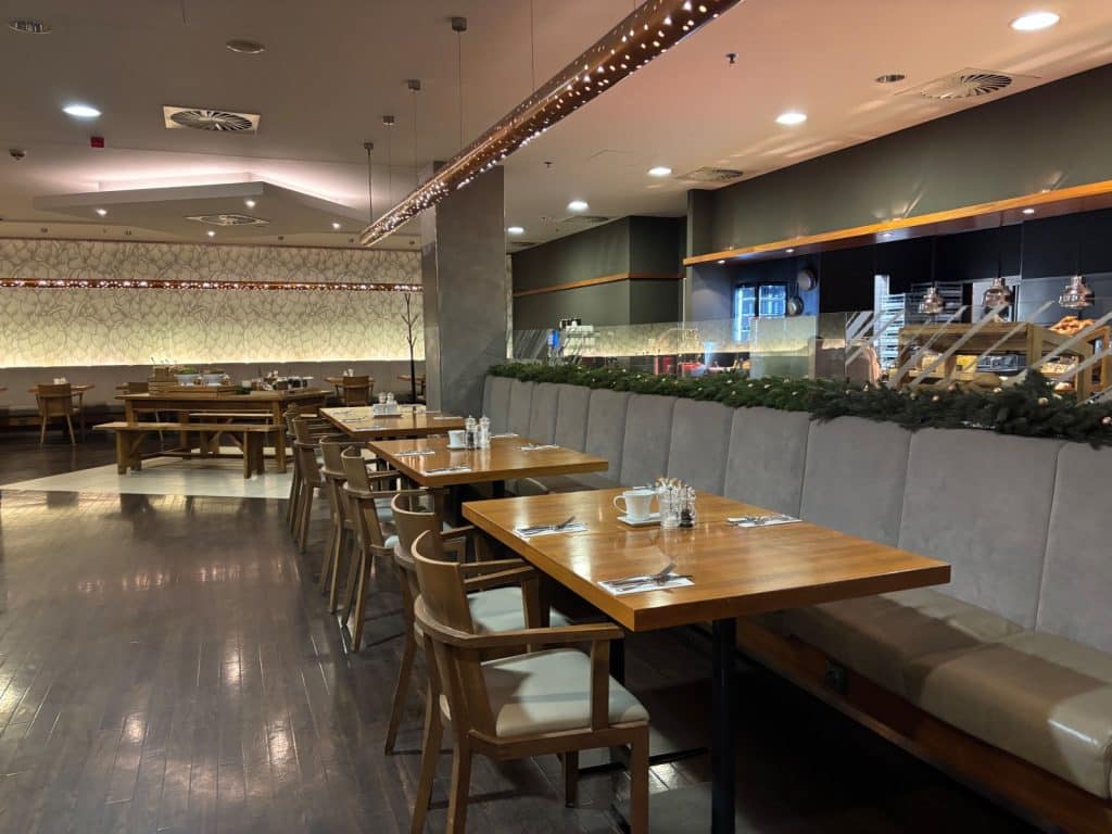 Das moderne Restaurant im Hotel zeigt eine Reihe von Holztischen mit grauen Polsterstühlen und einer langen Sitzbank vor einer mit Tannenzweigen dekorierten Glaswand. Im Hintergrund ist ein weitläufiger Speisesaal mit warmem Licht, einer gemusterten Wandgestaltung und einer offenen Buffetstation zu sehen.